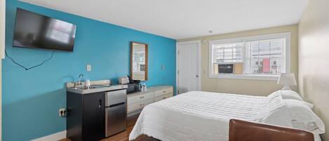 A hotel room with a bed, a desk, a kitchenette, a TV, and a window with blinds.