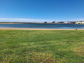Ein grünes Feld mit einem Gewässer im Hintergrund, eine Person sitzt im Gras, und Gebäude gegenüber dem Wasser.