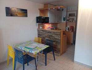 A dining area with a table and chairs, a kitchen with a microwave, and a painting on the wall. - Apartment 500m from the sea and 2min from Etel! (Erdeven)