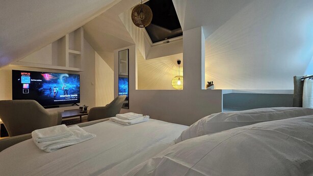 A hotel room with a bed, a desk with a TV displaying a Netflix interface, a chair, and a unique ceiling design.