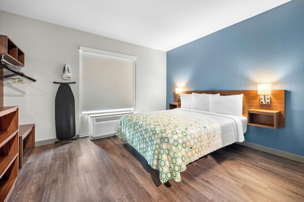 Habitación de hotel con cama, suelo de madera, ventana, calentador, estante con zapatos y lámpara fijada a la pared.