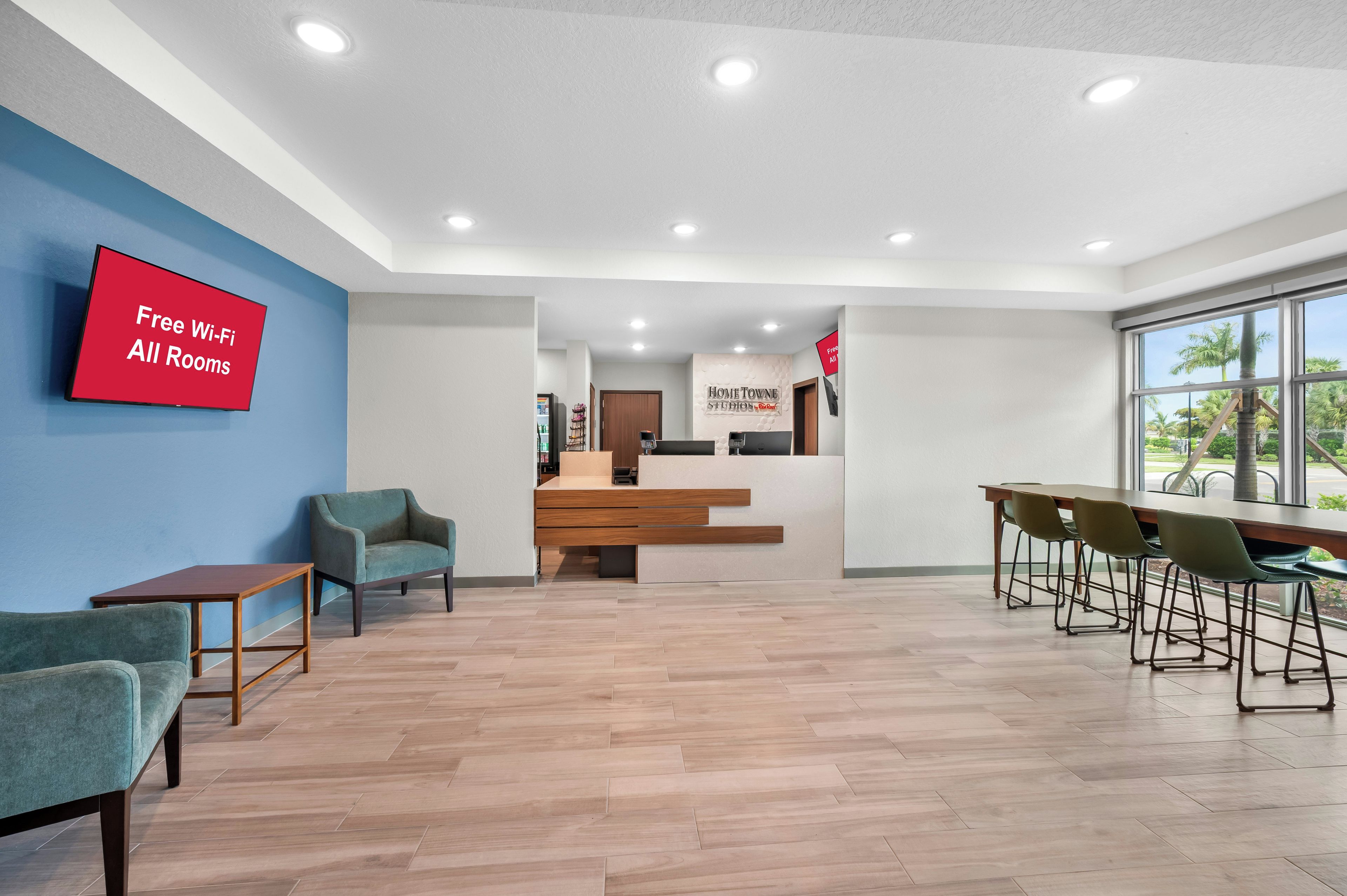 A hotel lobby with a reception desk, a TV displaying 'Free Wi-Fi All Rooms,' and a large window with a view of palm trees.