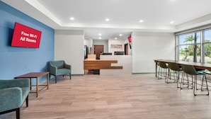 A hotel lobby with a reception desk, a TV displaying 'Free Wi-Fi All Rooms,' and a large window with a view of palm trees. - HomeTowne Studios by Red Roof Fort Myers (Fort Myers)