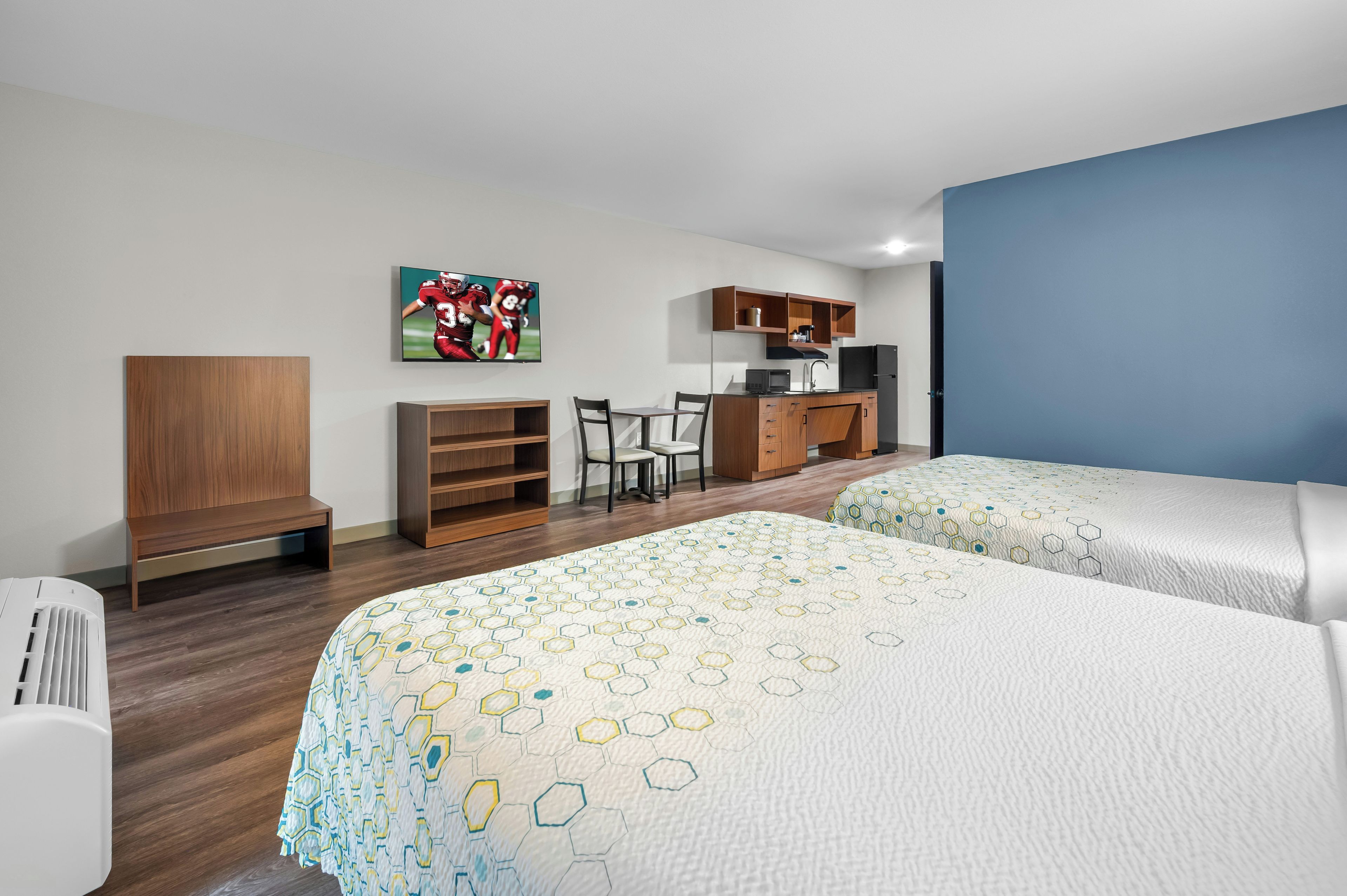 A hotel room with two beds, a desk, a chair, a TV, and a kitchenette.