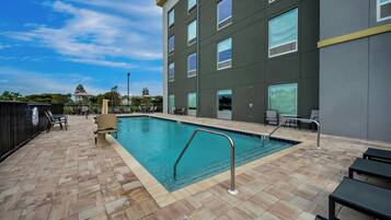 A swimming pool with a tiled deck, lounge chairs, and a building with multiple windows.