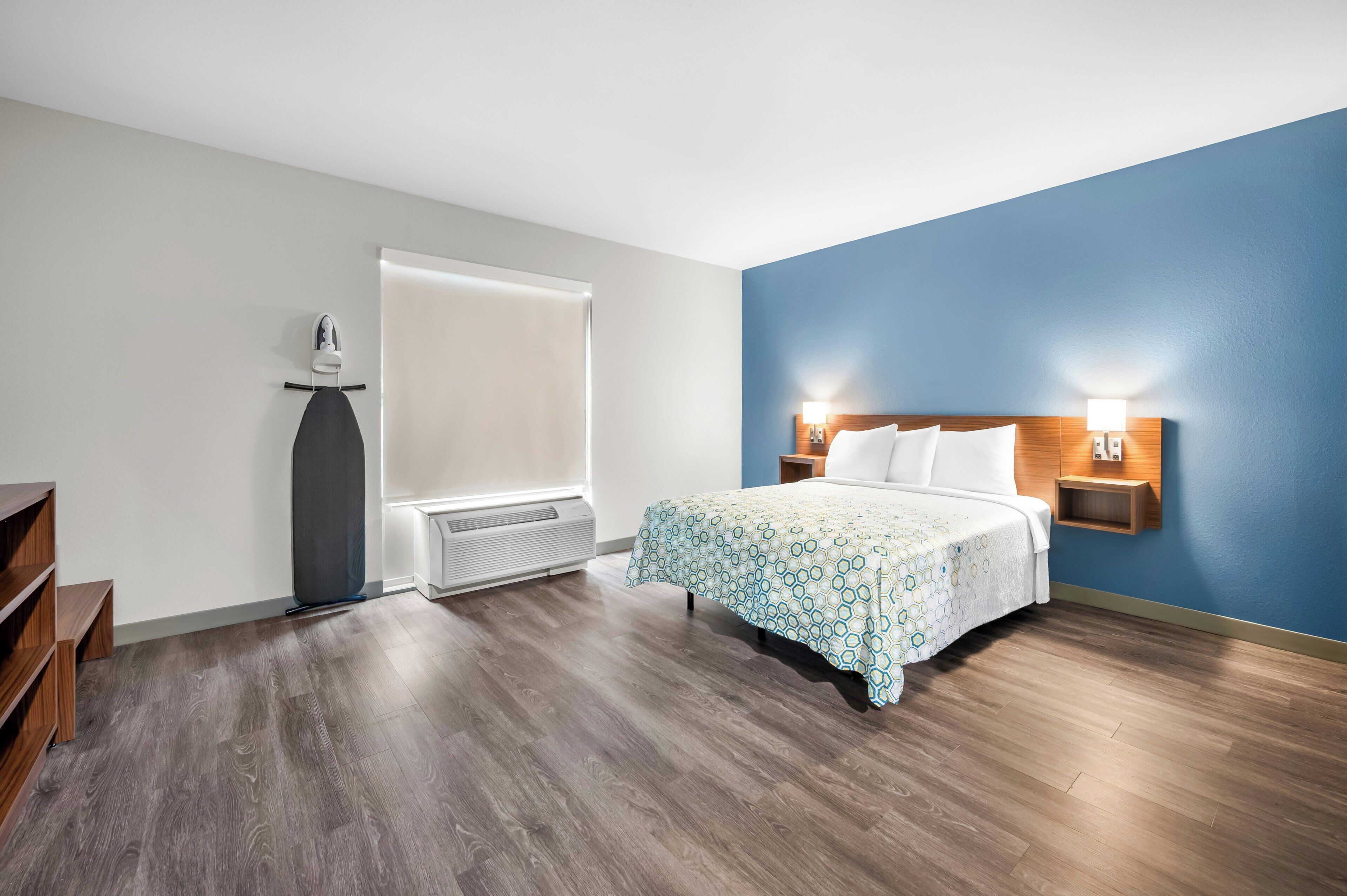 A hotel room with a bed, a heater, a window, and a wall-mounted nightstand.