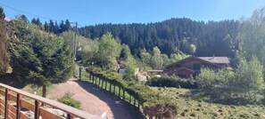 Paisaje rural con una valla de madera, un sendero de tierra y una casa rodeada de árboles y una colina boscosa.