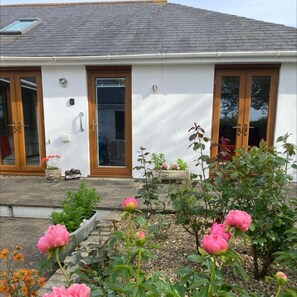A single-story house with a white exterior, a tiled roof, and a front porch with a small garden. - Honeysuckle Rise (Looe)