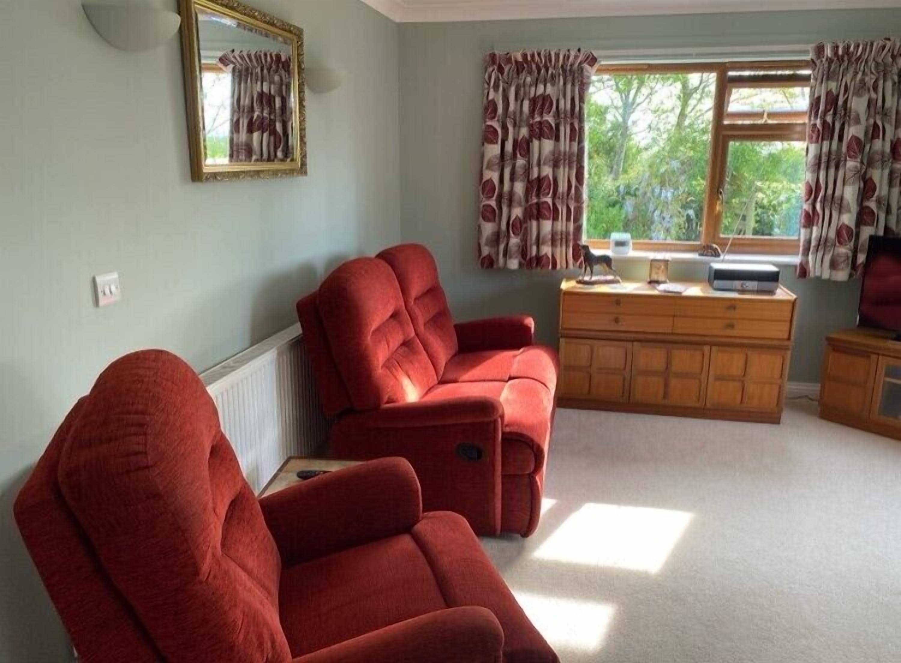 A room with two red armchairs, a wooden dresser, a mirror, and a window with curtains.