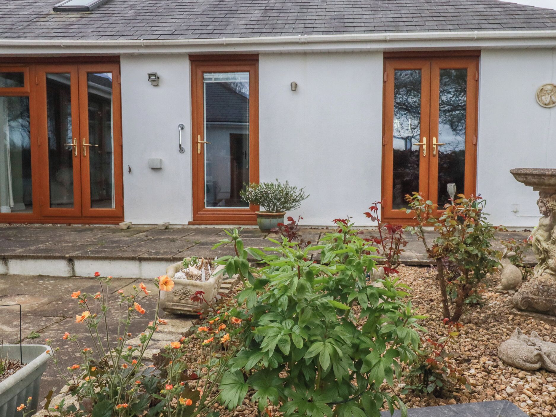 A white building with three wooden-framed glass doors, a small garden with various plants, and a stone sculpture.