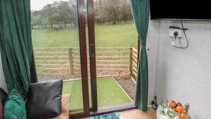 A room with a sliding glass door to a grassy outdoor area, a wooden bench, a sofa, and a table with a bottle and glasses. - The Horse Box (Blaydon on Tyne)