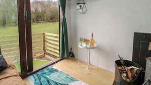 A room with a wooden floor, a bed, a TV mounted on the wall, a small table with a bottle and glasses, and a window with a view of a grassy area. - The Horse Box (Blaydon on Tyne)