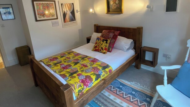 Une chambre à coucher avec un lit en bois, une couvre-lit colorée, une petite table de chevet, une lampe de chevet, une chaise et des œuvres d’art accrochées au mur.