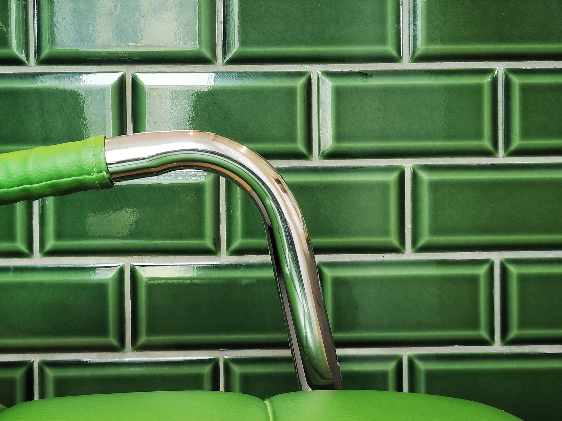 A green faucet with a chrome finish against a green tiled wall.