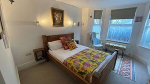 A bedroom with a wooden bed, a floral quilt, a chair, a table, and a window with blinds. - Church Retreat (St Leonards-On-Sea)