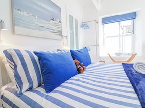 A bed with striped bedding, a teddy bear, and a window with a view of buildings. - Hideaway Cottage (Bideford)