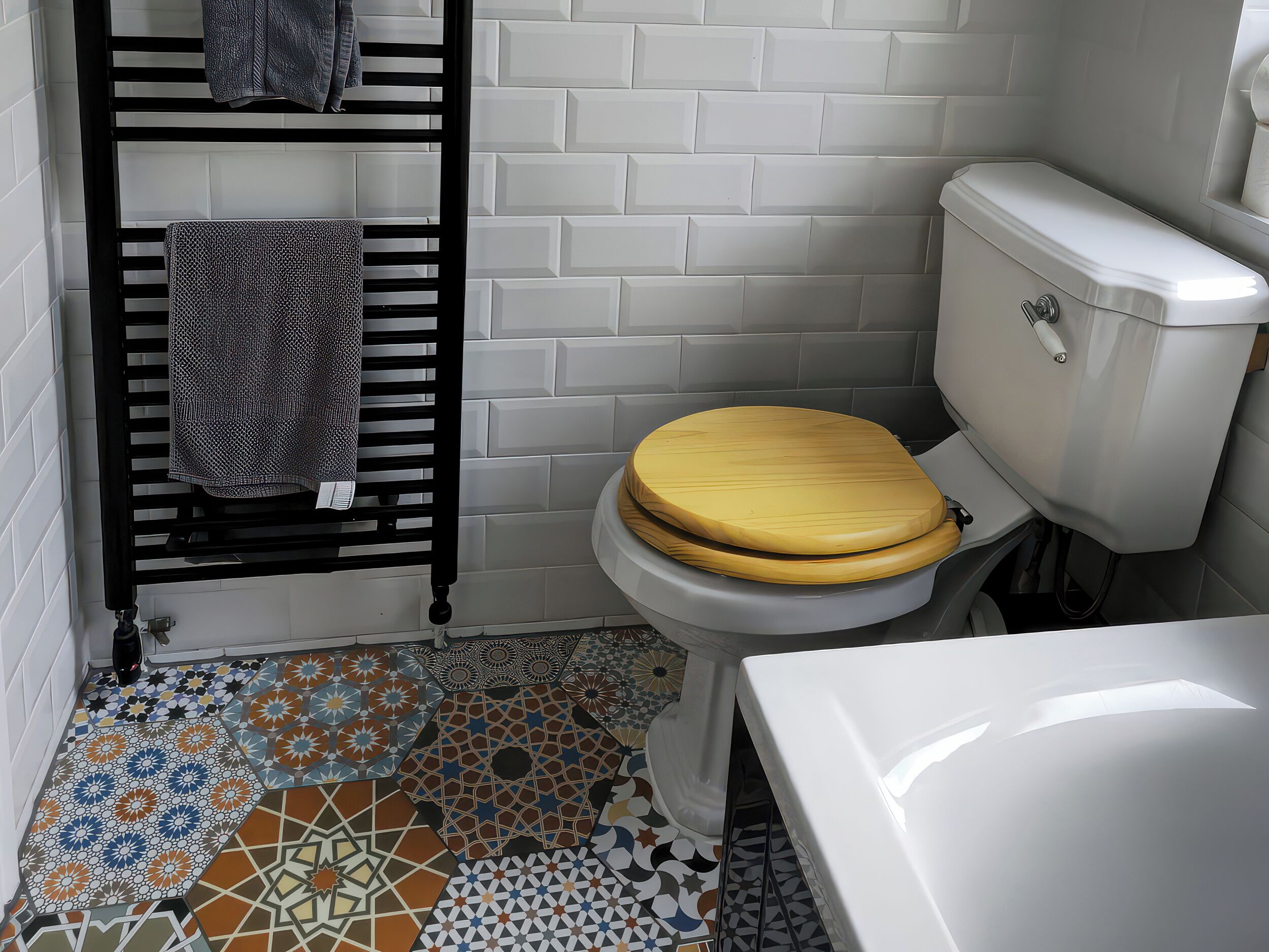 A bathroom with a white toilet, a wooden seat, a black towel rack, and a tiled floor with various patterns.