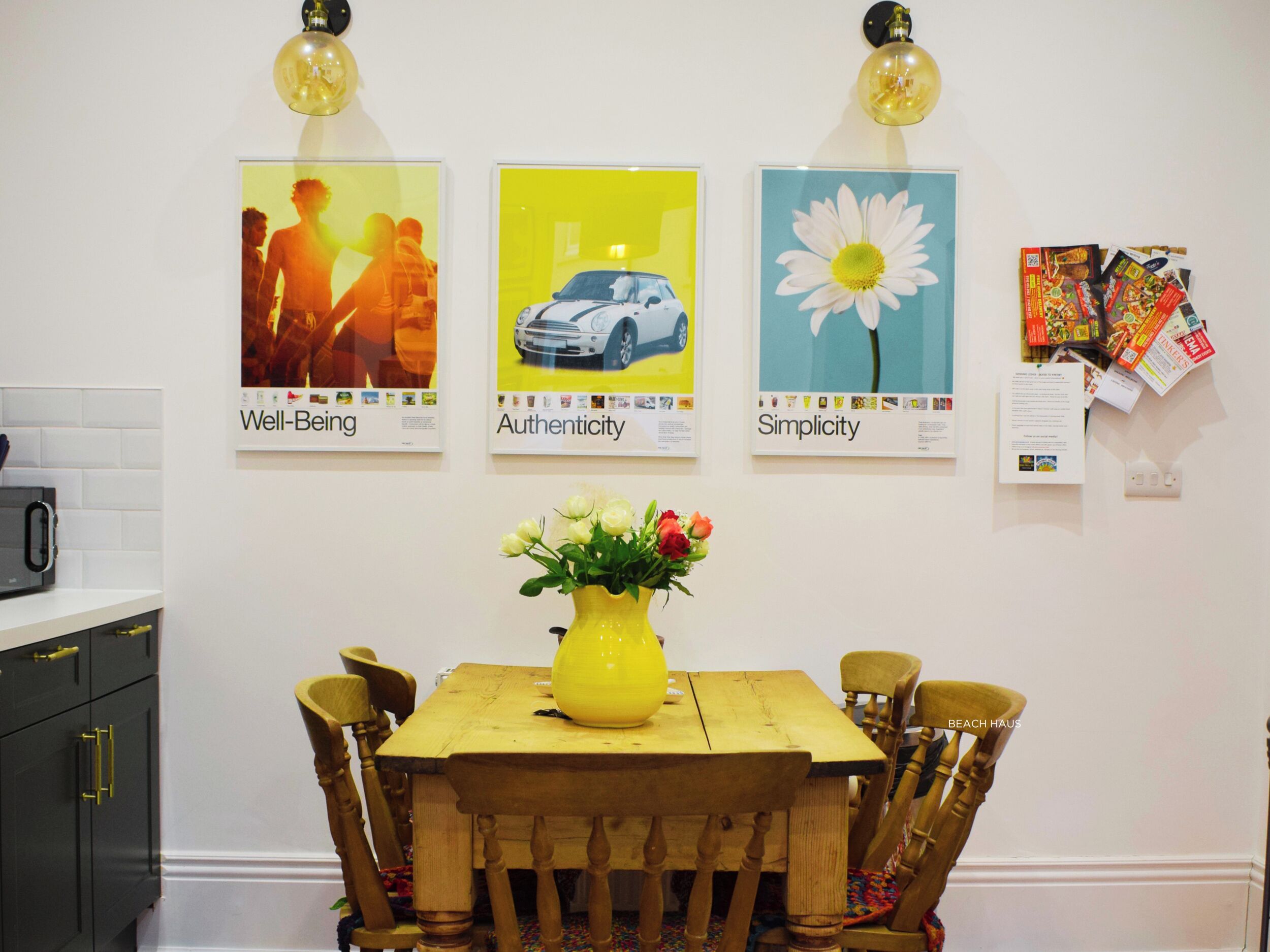 Un comedor con una mesa de madera, sillas y un jarrón amarillo con flores. Tres carteles enmarcados adornan la pared, cada uno con un tema diferente.