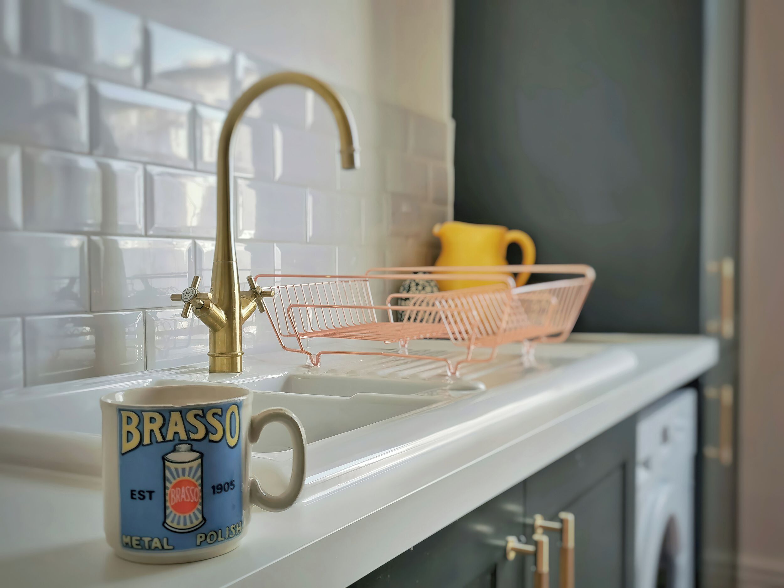 Eine Küche mit weißer Arbeitsplatte, einem Messing-Wasserhahn, einem pinken Drahtkorb und einer Tasse mit dem Aufdruck 'Brasso Metal Polish'.