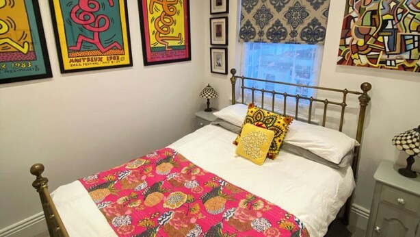 A bedroom with a bed, colorful artwork on the wall, a patterned window treatment, and a bedside table with a lamp. - 21 Gensing Road (St Leonards-On-Sea)