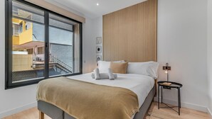 A modern bedroom with a large window, a bed with a beige blanket, a bedside table with a lamp, and a wooden panel wall. - Home2Book Modern & Cozy Las Damas (Las Palmas de Gran Canaria)