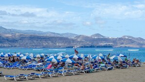A beach with rows of sun loungers and umbrellas, a clear sky, and mountains in the background. - Home2Book Modern & Cozy Las Damas (Las Palmas de Gran Canaria)