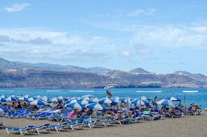 Ein Strand mit Reihen von Liegestühlen und Sonnenschirmen, ein klarer Himmel und Berge im Hintergrund.