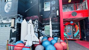 Un murale a tema spaziale con un navicella e pianeti, una facciata di edificio rosso e nero, e un ingresso con tappeto rosso.