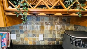 A kitchen with a tiled backsplash, a wooden shelf with hanging glassware, and a microwave.