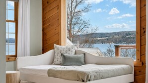 Een slaapkamer met een bed, een raam met gordijnen en uitzicht op een meer en heuvels.