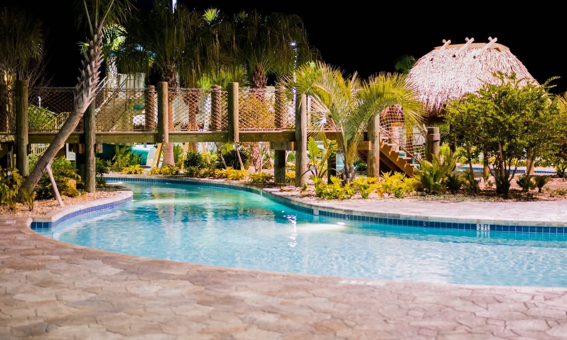 A swimming pool with a wooden deck, surrounded by tropical plants and a thatched-roof structure.