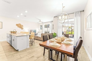 A modern kitchen with a central island, a dining area with a wooden table, and a living space with a sofa. - Bright Luxe At Championsgate (Davenport)