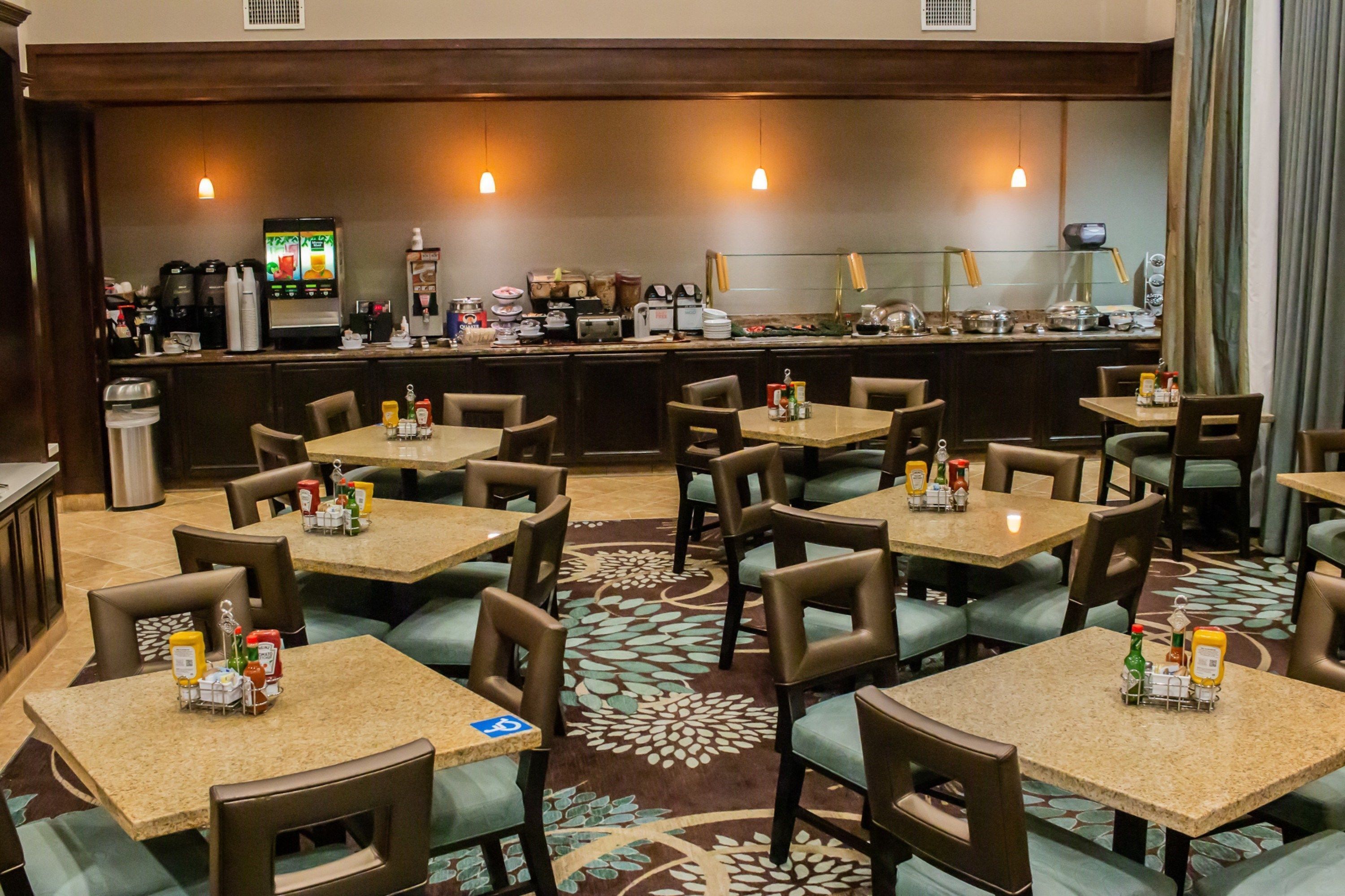 A restaurant with multiple square tables and chairs, a bar area with various appliances, and a patterned carpet.