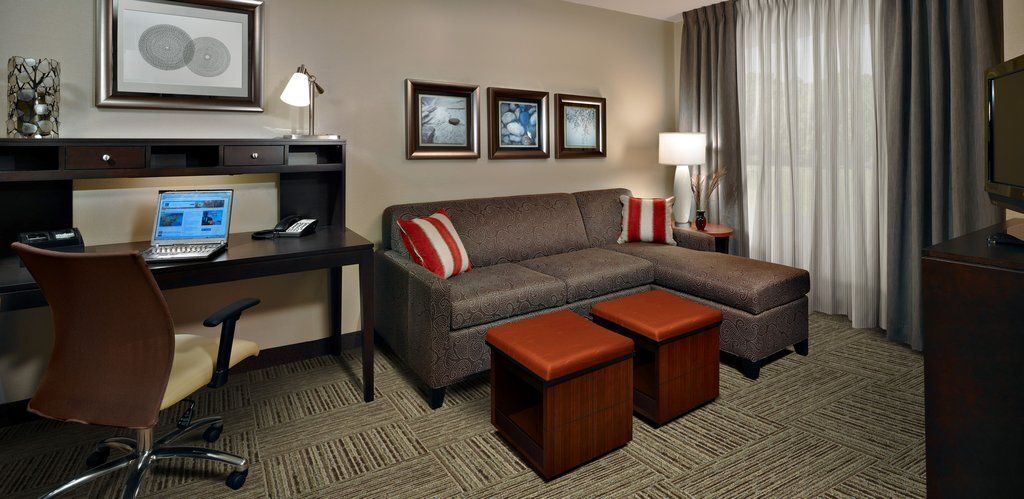 Habitación de hotel con escritorio, laptop, sillón de cuero marrón, sofá marrón con almohadas rojas y blancas, dos otomanas de madera, televisor y cuadros en la pared.
