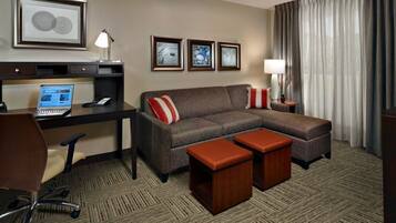 Habitación de hotel con escritorio, laptop, sillón de cuero marrón, sofá marrón con almohadas rojas y blancas, dos otomanas de madera, televisor y cuadros en la pared.