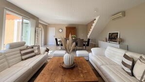 Un salon moderne avec une table basse en bois, des canapés blancs, un coin repas agrémenté d’un lustre et un escalier menant à un niveau supérieur.