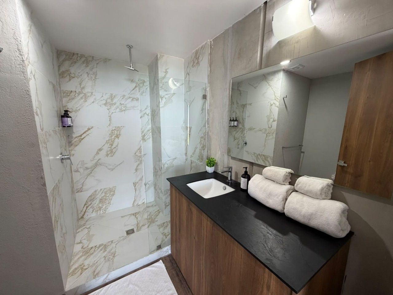 Une salle de bain avec un mur de douche en marbre, un meuble-lavabo en bois avec un plan de travail noir et un miroir au-dessus du lavabo.