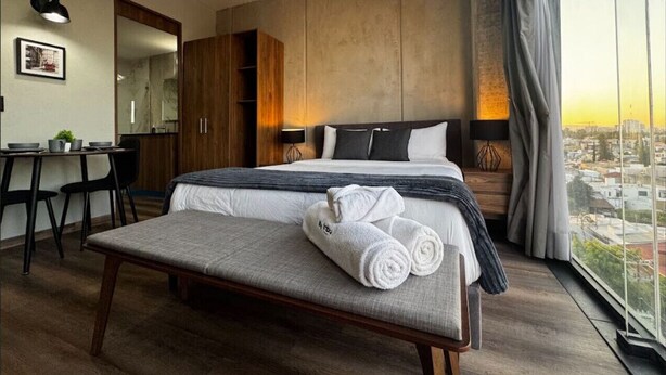 A modern hotel room with a bed, a dining table, a bench, and a large window with a city view. - Amazing Suite consulado (Guadalajara)
