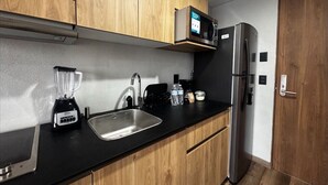 A compact kitchen with wooden cabinets, a black countertop, a stainless steel refrigerator, a microwave, a blender, and a sink. - Amazing Suite consulado (Guadalajara)