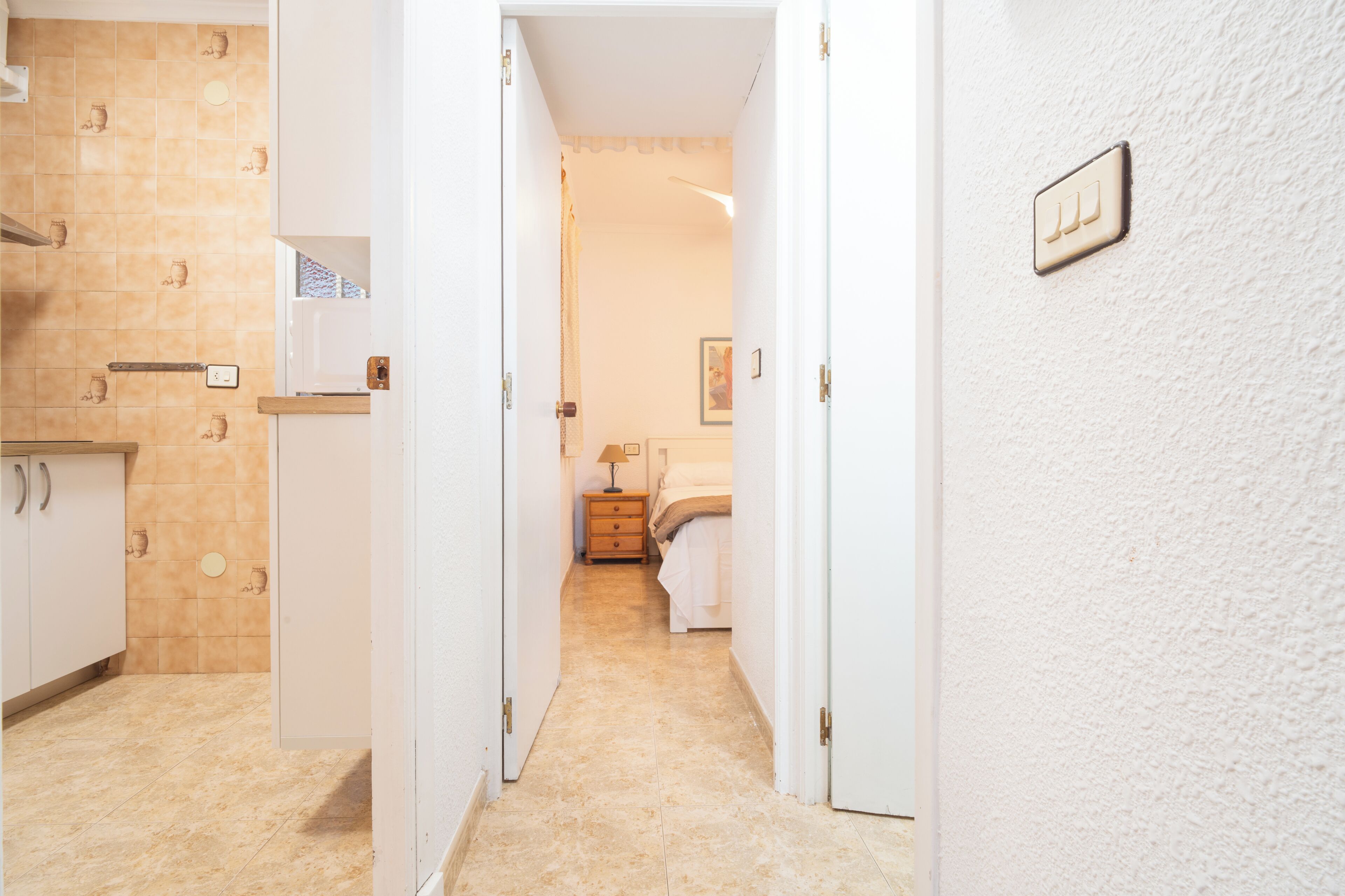 Ein Flur mit einer Küche links, einem Schlafzimmer mit Bett und Nachttisch in der Mitte und einer weißen Wand mit Lichtschalter rechts.