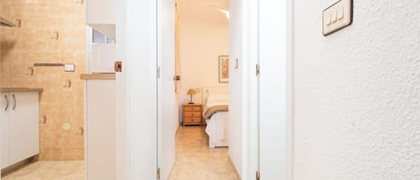 A hallway with a kitchen on the left, a bedroom with a bed and nightstand in the center, and a white wall with a light switch on the right.