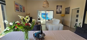 A living room with a white sofa, a dining table with a vase of flowers, a bottle of champagne, and a view of a pool through glass doors. - Villa with indoor pool 150m from the beach (Plounéour-Brignogan-plages)