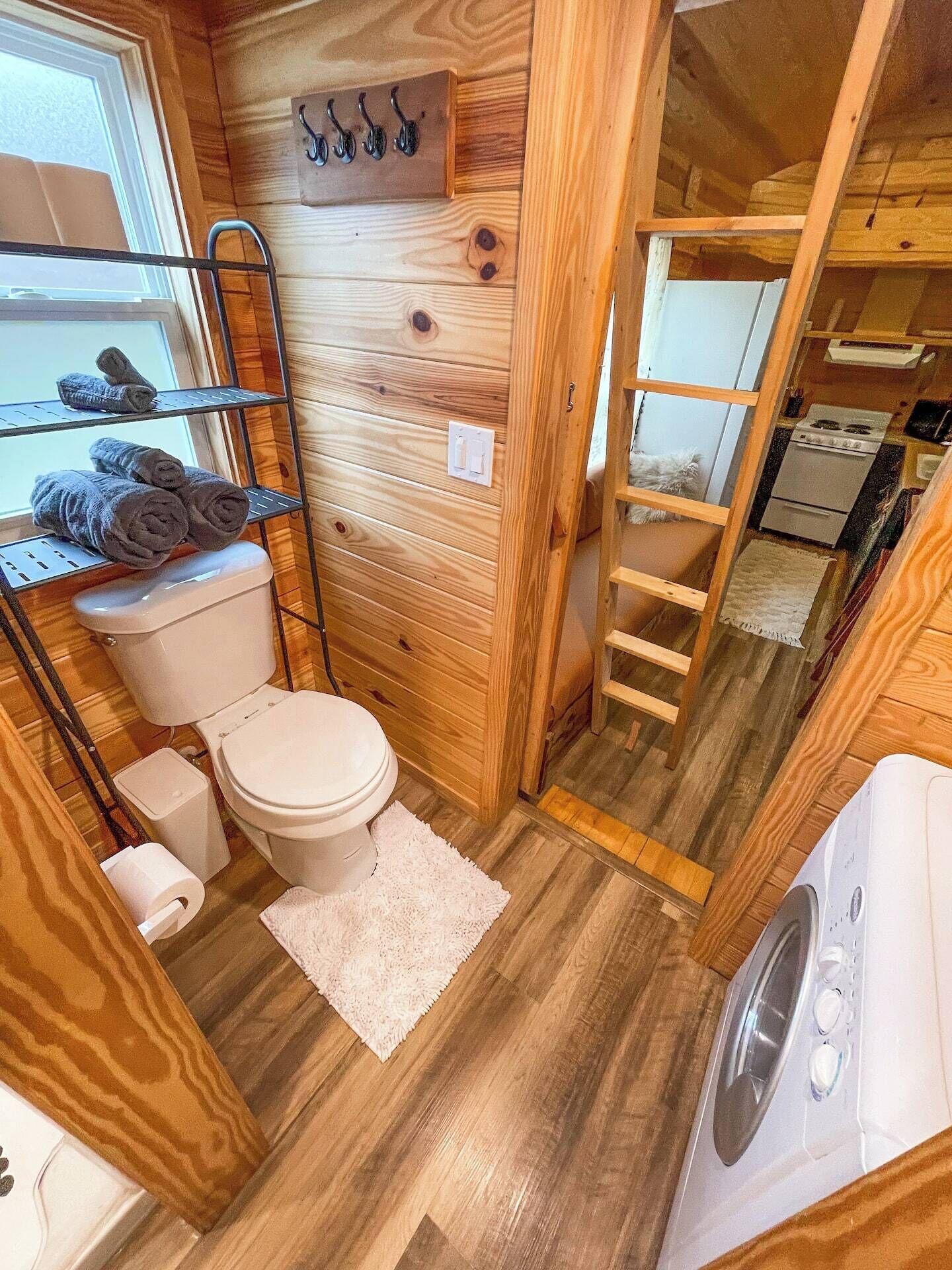 A compact wooden bathroom with a toilet, a shelf with towels, a small sink, and a washing machine.