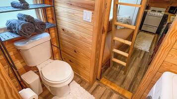 A compact wooden bathroom with a toilet, a shelf with towels, a small sink, and a washing machine.