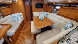 Interior de un yate con cubierta de madera, una zona de estar central con una mesa y un televisor montado en la pared.