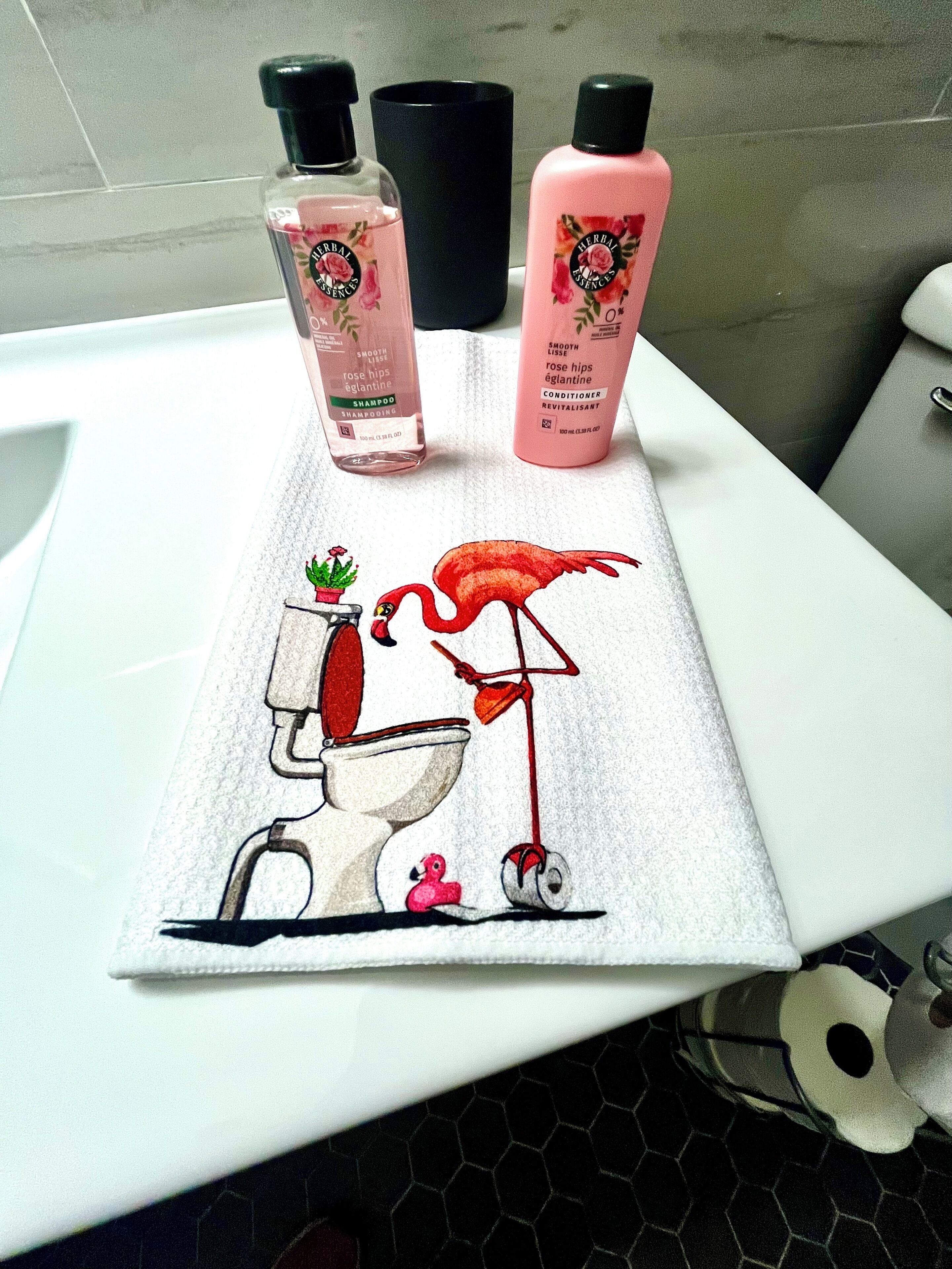 A bathroom with a white sink, two bottles of rose hips essence shampoo and conditioner, and a towel with a toilet and flamingo illustration.