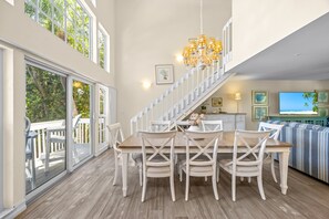A spacious dining area with a wooden table and white chairs, a staircase leading to an upper level, and a view of the outdoors through large windows. - Slip into island time the moment you arrive at Poppy’s Paradise (Captiva)