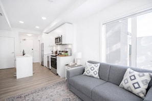 A modern living room with a grey sofa, a small side table, and a kitchen area in the background. - New Suite • Near DT Stampede• UG Park • Banff Pass (Calgary)