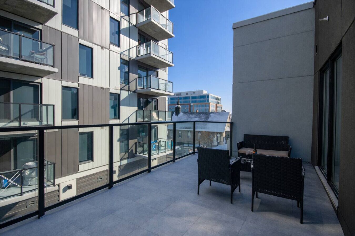 Un balcón con mobiliario exterior, barandilla de vidrio y vistas a edificios cercanos.
