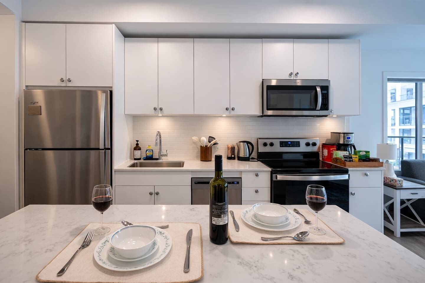 A modern kitchen with stainless steel appliances, white cabinetry, and a dining area set for two with wine glasses and a bottle.